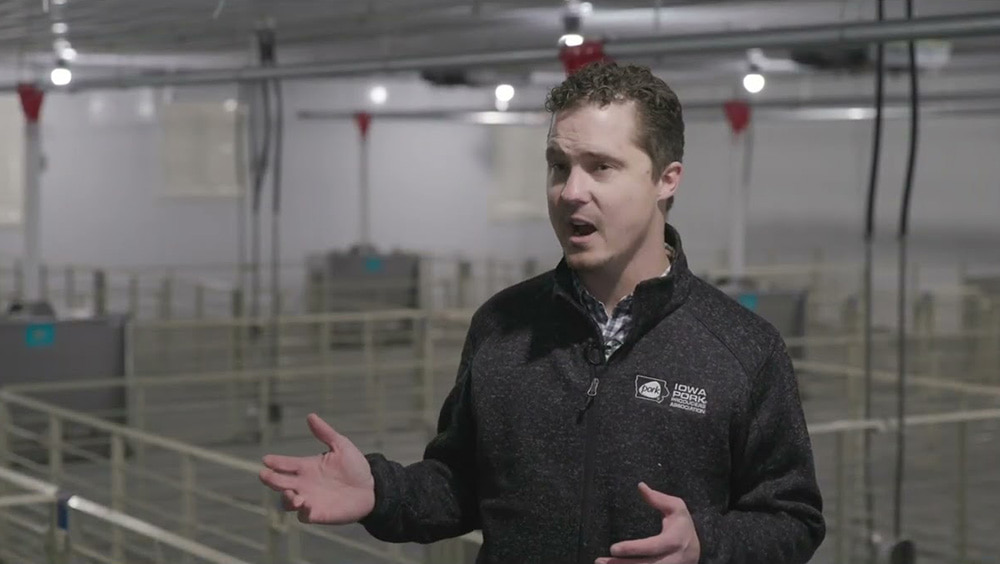 A man in a dark jacket with an 'Iowa Pork Association' logo is speaking with both hands raised, standing inside a modern indoor facility with metal railings, overhead lights, and equipment in the background.